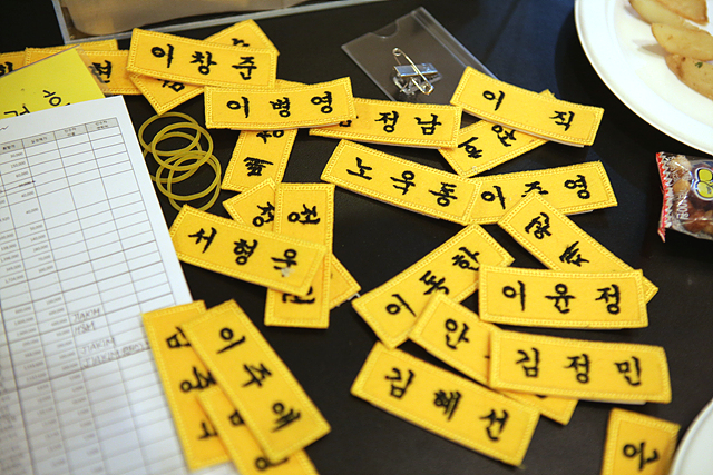 노란색 천 이름표에 검정색 실로 이름이 자수로 새겨져 있고, 여러 개의 이름표들이 테이블 위에 어지럽게 놓여 있다.