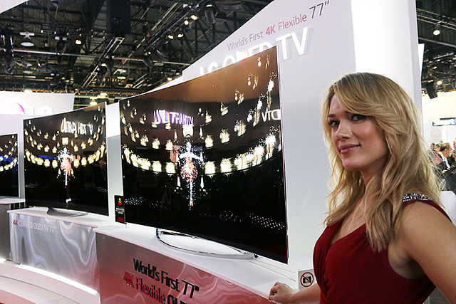 금발의 여자 모델이 가변형 UHD 올레드 TV 앞에 서있다. 옆으로 곡선으로 휘어진 TV두 대가 나란히 놓여있다.