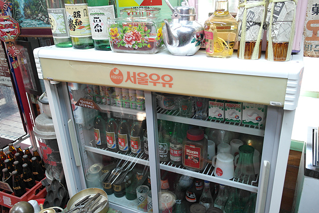 한국 근현대사 박물관에 전시되어 있는 옛날 우유냉장고. 냉장고 안쪽에 옛날 우유와 술병이 들어 있고 우유냉장고 위에 양은주전자 등 옛날 소품들이 놓여 있다.