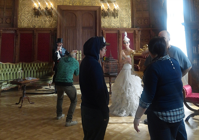흰 드레스를 입은 모델과 턱시도를 입은 모델이 촬영을 준비하고 있으며 그 앞으로 스태프들이 대화를 나누고 있다.