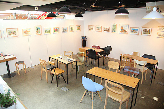 샐프 전시회를 열 수 있는 갤러리 내부전경, 하얀 벽에 다양한 작품들이 걸린 액자가 걸려있고 가운데 책상과 의자들이 놓여져 있다