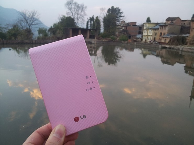 배경으로 네팔의 호수가 보이고 멀리 집과 나무들이 보인다. 2014년 포켓포토 분홍색 제품이 앞쪽에 크게 보인다.