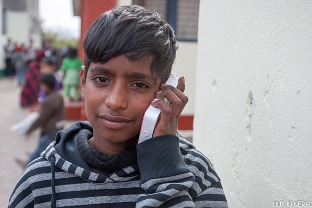 네팔 남학생이 포켓포토를 한 손에 들고 귀에 가까이 대어 사진이 나오고 있는 소리를 듣고 있다.
