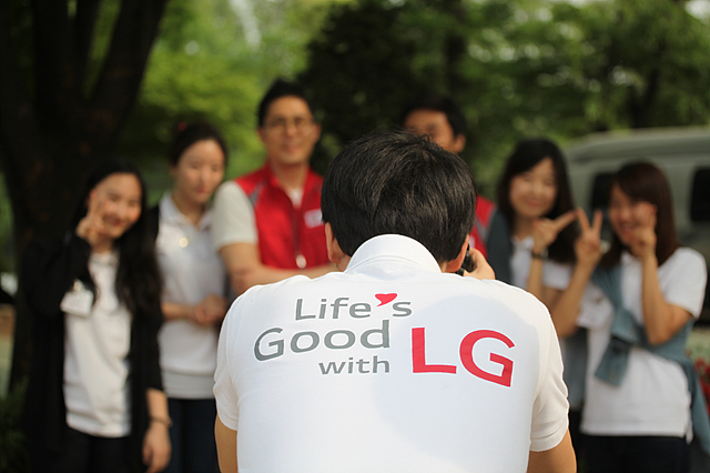 Life’s Good With LG’ 라고 쓰여진 흰 티셔츠사내가 사람들의 단체사진을 찍어주고 있다