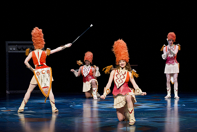 기다란 주황색 털모자를 쓰고 악대 같은 옷을 입은 배우 네명이 무대에서 춤을추고 있는 모습이다