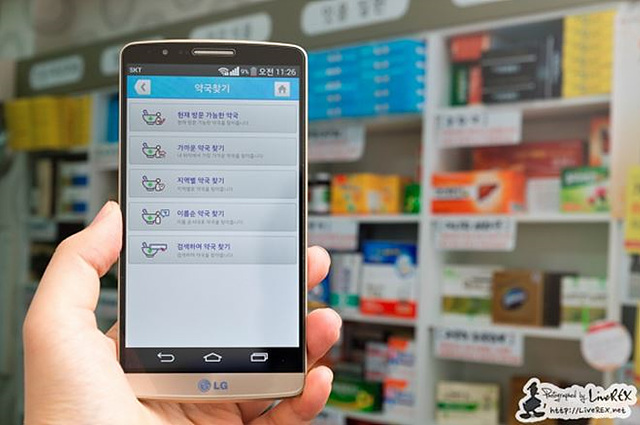 약국을 배경으로 약국찾기 어플리케이션 페이지가 구동되는 스마트폰을 한 손으로 들고 있다.