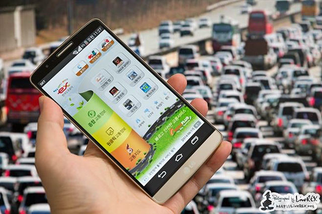 교통 체증이 심각한 도로를 배경으로 고속도로교통정보 어플리케이션이 구동되는 스마트폰을 한 손으로 들고 있다.