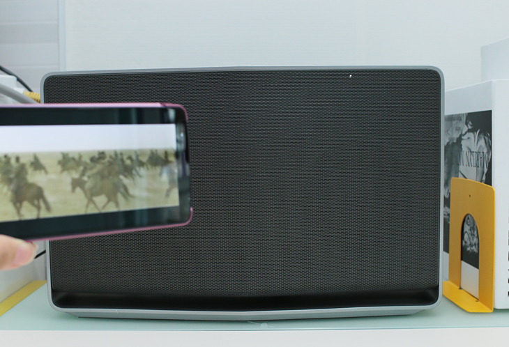 블루투스를 통해 스마트폰으로 음악을 재생하고 있다. 스마트폰 뒤에는 탁상 위에 놓여 있는 LG 스마트 오디오가 보인다. 