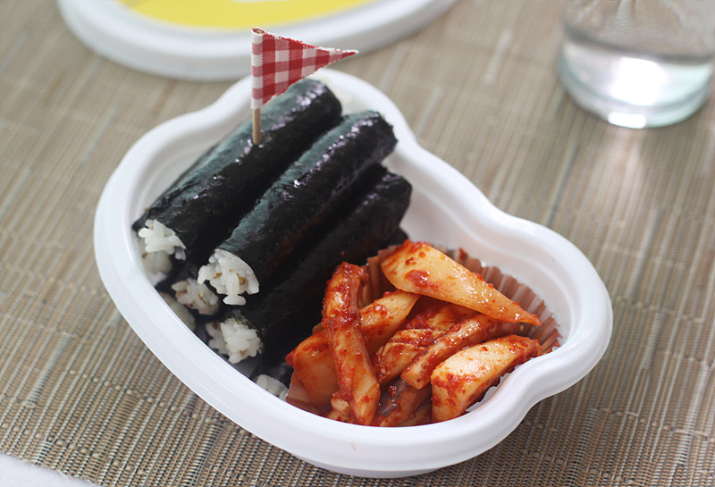 깃발 모양의 이쑤시개가 꽂혀있는 충무김밥. 매콤한 양념이 된 오징어가 함께 놓여있다.