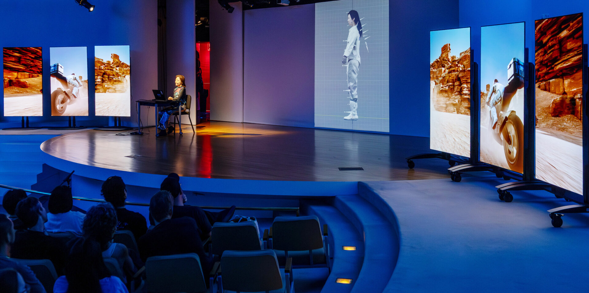 넓은 무대 위에서 한 사람이 노트북 앞에 앉아 시연을 진행하고 있습니다. 무대 좌우에는 세로로 긴 대형 화면들이 설치되어 있으며, 오토바이를 타고 사막을 달리는 장면과 붉은 바위 협곡 풍경이 생생하게 재생되고 있습니다. 중앙 스크린에는 흰색 복장을 한 인물이 측면에서 서 있는 3D 스캔 이미지가 표시되어 있습니다. 객석에는 여러 관객이 앉아 무대를 주목하고 있습니다.