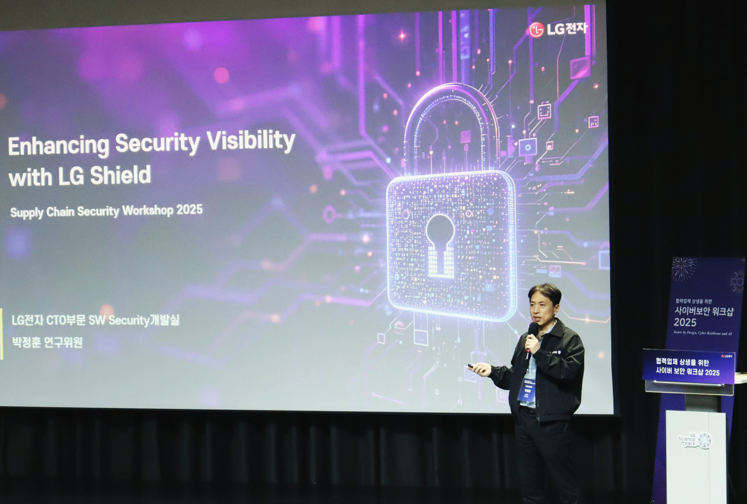 무대 위 스크린에 보라색 배경으로 자물쇠 아이콘이 크게 표시되어 있으며, ‘Enhancing Security Visibility with LG Shield’라는 제목이 영어로 쓰여 있다. 하단에는 ‘LG전자 CTO부문 SW Security개발실 박정훈 연구위원’ 문구가 있다. 오른쪽에는 박정훈 연구위원이 마이크를 들고 발표 중이다.